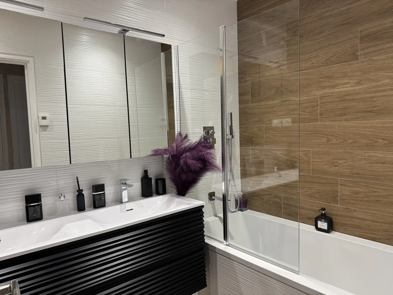 Aménagement de salle de bains moderne à Orsay, avec une vasque en Corian élégante aux contours noirs, murs habillés de carreaux blancs à effet bois, et paroi vitrée transparente pour une ambiance lumineuse et contemporaine.