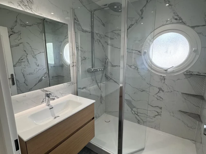 Rénovation de salle de bains à Bondoufle alliant élégance et modernité : murs en marbre, douche à l’italienne spacieuse, vasque avec contour en bois et tiroir de rangement pratique, miroir éclairé, et paroi vitrée pour un espace lumineux et design.