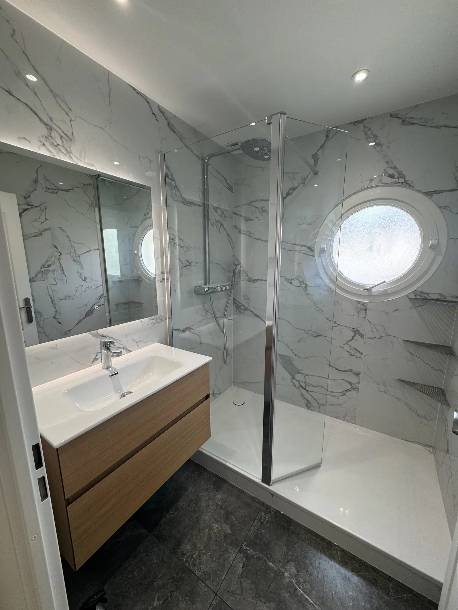 Rénovation de salle de bains à Bondoufle alliant élégance et modernité : murs en marbre, douche à l’italienne spacieuse, vasque avec contour en bois et tiroir de rangement pratique, miroir éclairé, et paroi vitrée pour un espace lumineux et design.