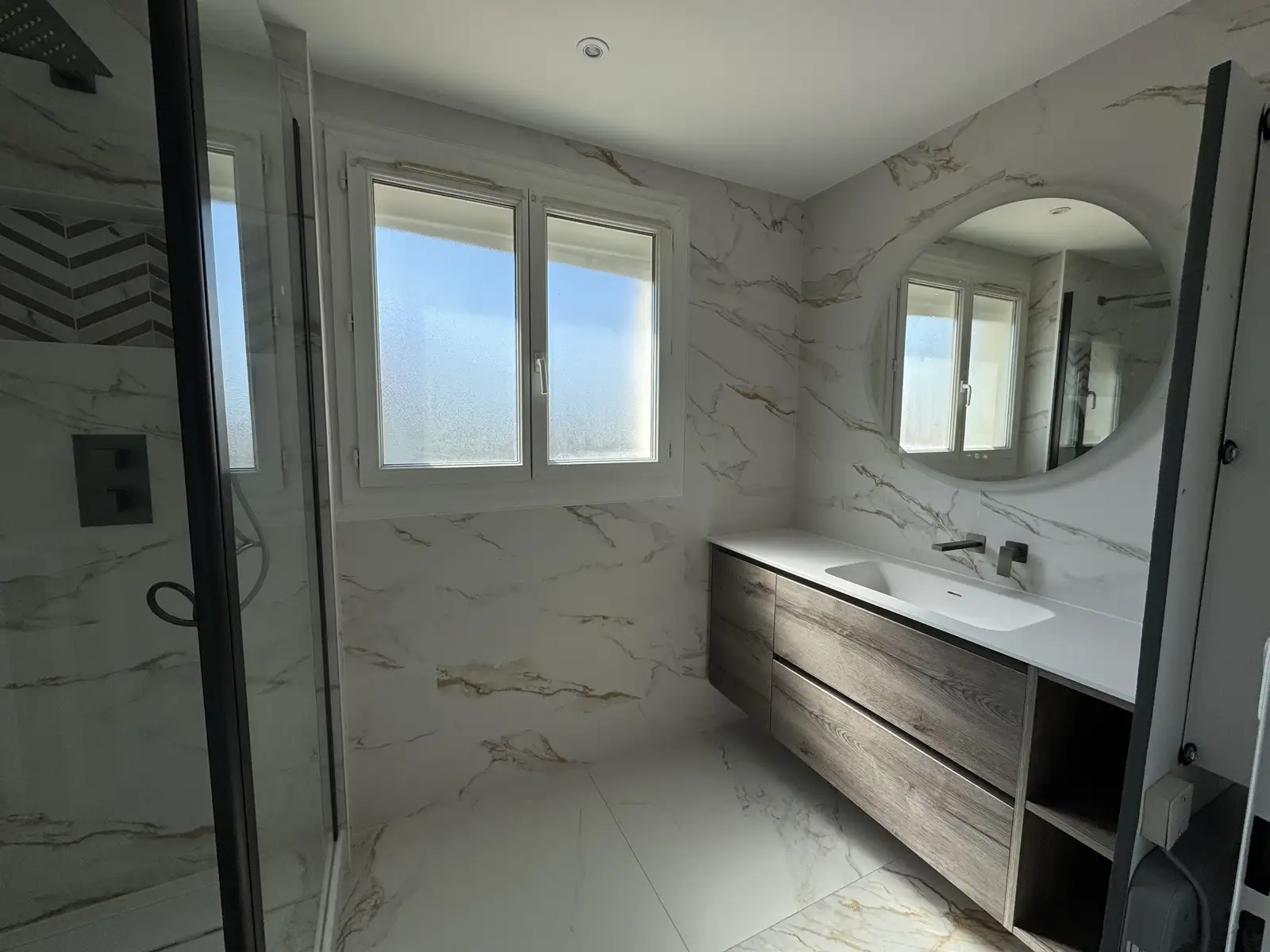 Salle de bains en marbre avec miroir arrondi suspendu et éclairé, vasque avec revêtement bois, tiroirs de rangement et placard